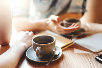 Auf einem Tisch sind Kaffeetassen, Schreibblöcke. Personen gestikulieren.